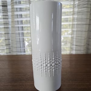 Puede incluir: Un jarrón alto de cerámica blanca con forma cilíndrica. La parte inferior presenta un patrón texturizado de puntos redondeados en relieve. El jarrón se exhibe sobre una superficie de madera oscura, con una cortina transparente al fondo. Aproximadamente 25 cm de alto.