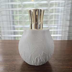 May include: A white fragrance lamp with a rounded, bulbous body featuring an embossed botanical pattern of leaves and stems. The top of the lamp is fitted with a polished gold-tone metal crown cap with vertical cutouts for ventilation.