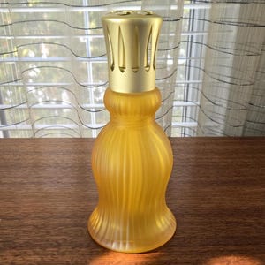 Può includere: Una lampada profumata in vetro giallo con un top in metallo color oro. La lampada ha una base arrotondata e un design strutturato e a coste. La parte superiore presenta fessure verticali per la diffusione della fragranza. La lampada è progettata per contenere oli profumati.