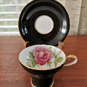 Könnte beinhalten: Ein schwarzes und goldenes Tee-Set mit einem rosafarbenen Rosenblütenmuster. Die Teetasse hat einen breiten, ausgestellten Boden und einen zarten Henkel.