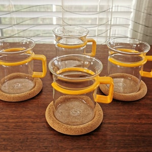 May include: Four clear glass mugs with yellow bands around the top and cork coasters. The mugs have rounded handles.