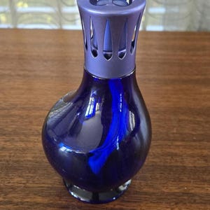 Pu&ograve; includere: Una bottiglia diffusore per aromaterapia in vetro blu scuro con una parte superiore decorativa e perforata. La bottiglia ha una forma arrotondata con una base scura. La parte superiore &egrave; di una tonalit&agrave; di blu pi&ugrave; chiara. La bottiglia &egrave; su una superficie di legno.