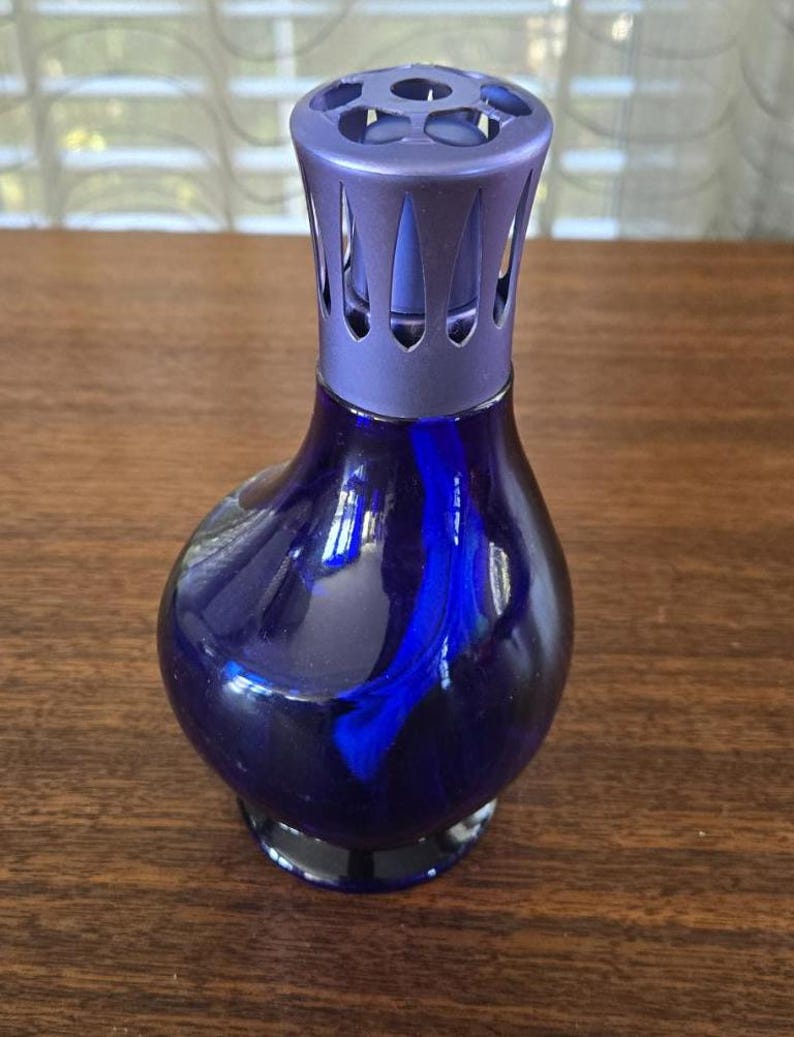 Pu&ograve; includere: Una bottiglia diffusore per aromaterapia in vetro blu scuro con una parte superiore decorativa e perforata. La bottiglia ha una forma arrotondata con una base scura. La parte superiore &egrave; di una tonalit&agrave; di blu pi&ugrave; chiara. La bottiglia &egrave; su una superficie di legno.