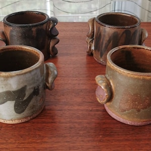 May include: Four ceramic mugs with unique brown and grey glaze patterns and sculpted handles. The mugs are handmade and have a rustic, earthy aesthetic.