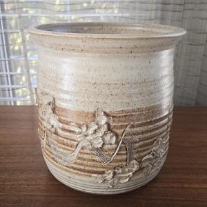 May include: A beige ceramic vase with a wide opening and a textured surface. The vase features a band of carved floral and linear designs in a darker brown hue. The overall aesthetic is rustic and handcrafted, suitable for home decor.