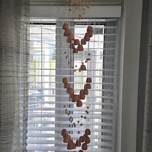 May include: A hanging mobile with a natural, earthy aesthetic. The mobile features small, terracotta-colored bell-shaped elements and wooden beads suspended from a woven, bowl-shaped top. The design is displayed in front of a window with white blinds and sheer curtains.