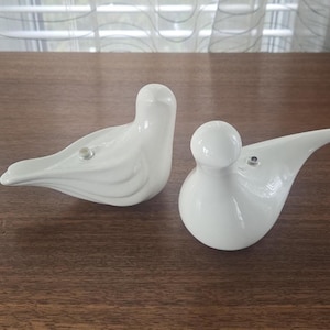 May include: Two white ceramic bird ornaments. One bird is upright, the other is resting. Both have a smooth, glossy finish and are on a dark wooden surface. The ornaments are decorative and add a touch of elegance to any room. The birds are decorative.