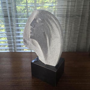 Op de afbeelding: Een lichtgrijze stenen sculptuur van een vrouwenprofiel, gemonteerd op een donkerbruine rechthoekige basis. De sculptuur heeft een getextureerd oppervlak en vloeiend haar. Het kunstwerk wordt tentoongesteld op een houten oppervlak.