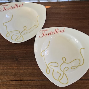 Op de afbeelding: Twee gebroken witte, driehoekige pastaborden. Het woord "Tortellini" staat in rood schrift bovenaan elke kom. Een goudkleurig, abstract pastadesign staat op de onderste helft van elke kom. De kommen staan op een donker houten oppervlak.