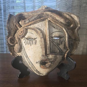 Puede incluir: Escultura de rostro de cerámica con ojos cerrados y rasgos faciales detallados. La escultura es en tonos marrones y beige, con líneas grabadas y una superficie texturizada. Está montada sobre un soporte negro, mostrando una pieza artística y artesanal.