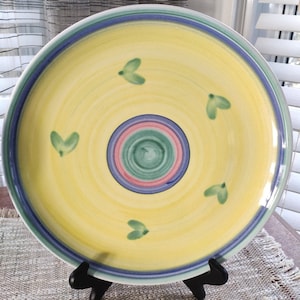 May include: A large yellow ceramic plate with a blue, green, and pink concentric circle design in the center. The plate has a blue and green rim and five green leaf-shaped designs around the center circle.