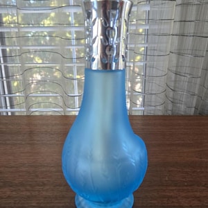 May include: A light blue frosted glass fragrance lamp with a silver metal decorative top. The lamp has a rounded body with a flared base and is decorated with a leaf pattern. The silver top has a decorative cut-out design.