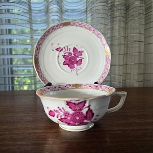 May include: A white teacup and saucer set with a floral design in shades of pink and gold trim. The teacup has a small handle and sits on a matching saucer. The set is placed on a dark wooden surface.