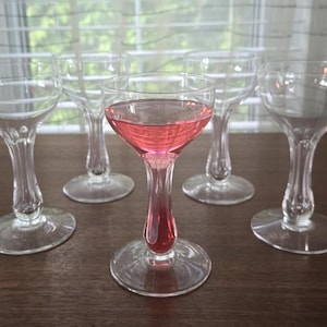 May include: Set of six clear glass stemmed cocktail glasses, one filled with pink liquid. The glasses have a delicate, fluted design.