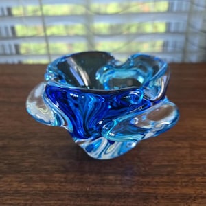 Puede incluir: Un cenicero de vidrio azul y transparente con un diseño ondulado y abstracto. El cenicero está hecho de vidrio soplado a mano y tiene una forma única y orgánica.