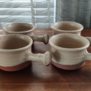 May include: Set of four stoneware soup bowls with handles. The bowls are a light beige color with a brown rim.