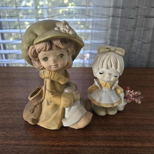 May include: Two ceramic figurines of children. One wears a large green hat and coat, holding a bundle. The other wears a yellow dress and holds a small bouquet of dried flowers. Both have detailed facial features.