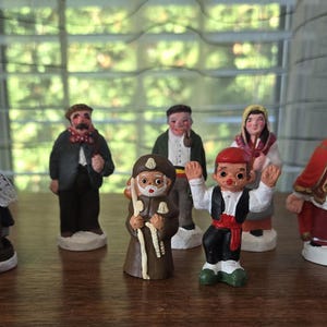 May include: A collection of small, hand-painted figurines depicting various figures in traditional attire. The figurines include an elderly woman, a man in a suit, and a figure resembling Saint Nicholas, all standing on a wooden surface.