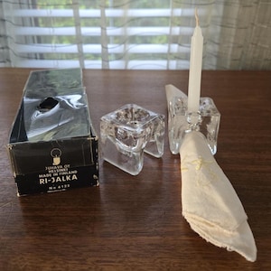 May include: Clear glass candle holders with a white candle and a folded napkin. The candle holders are square and have a unique design. A black box with text is also present. The text on the box reads "JUHAVA OY HELSINKI MADE IN FINLAND RI-JALKA No 6122".