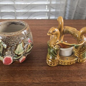 May include: Two ceramic candle holders shaped like geese. The geese are brown, green, and yellow with a white candle in the center. The candle holders are on a wooden surface.