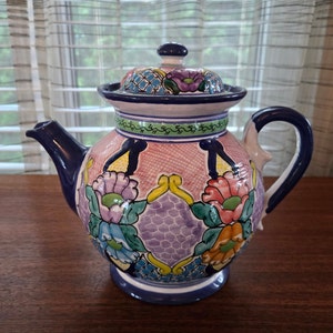 May include: A hand-painted ceramic teapot with a lid. The teapot features a pink body with floral designs in shades of purple, blue, green, and orange. The spout, handle, and base are dark blue, and the lid has a decorative pattern.