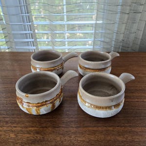 May include: Four ceramic soup bowls with handles. The bowls are beige with a brown and orange stripe around the middle and a white base. The bowls are arranged on a wooden surface.