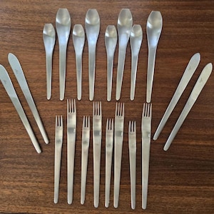 May include: A collection of stainless steel flatware, including spoons, forks, and knives, arranged on a wooden surface. The spoons have an oval bowl shape, and the knives have a long, slender design. The forks have three or four tines.