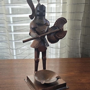 May include: A metal knight figurine holding a shield and axe, standing on a square base with a round ashtray. The knight is wearing armor and a helmet with a plume.