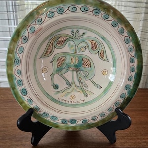 May include: A decorative ceramic plate with a hand-painted design. The plate features a central image of a stylized animal under a tree, with green, blue, and brown accents. The rim has a green border with a swirl pattern. The plate is displayed on a black stand.