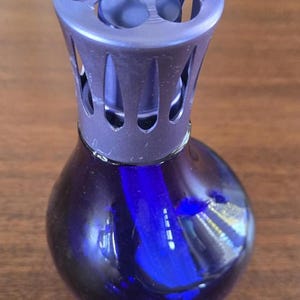 Pu&ograve; includere: Una bottiglia diffusore di fragranze in vetro blu scuro con un tappo decorativo, traforato, blu-viola. La bottiglia ha una forma arrotondata e una superficie liscia e riflettente. Il tappo ha un design geometrico.