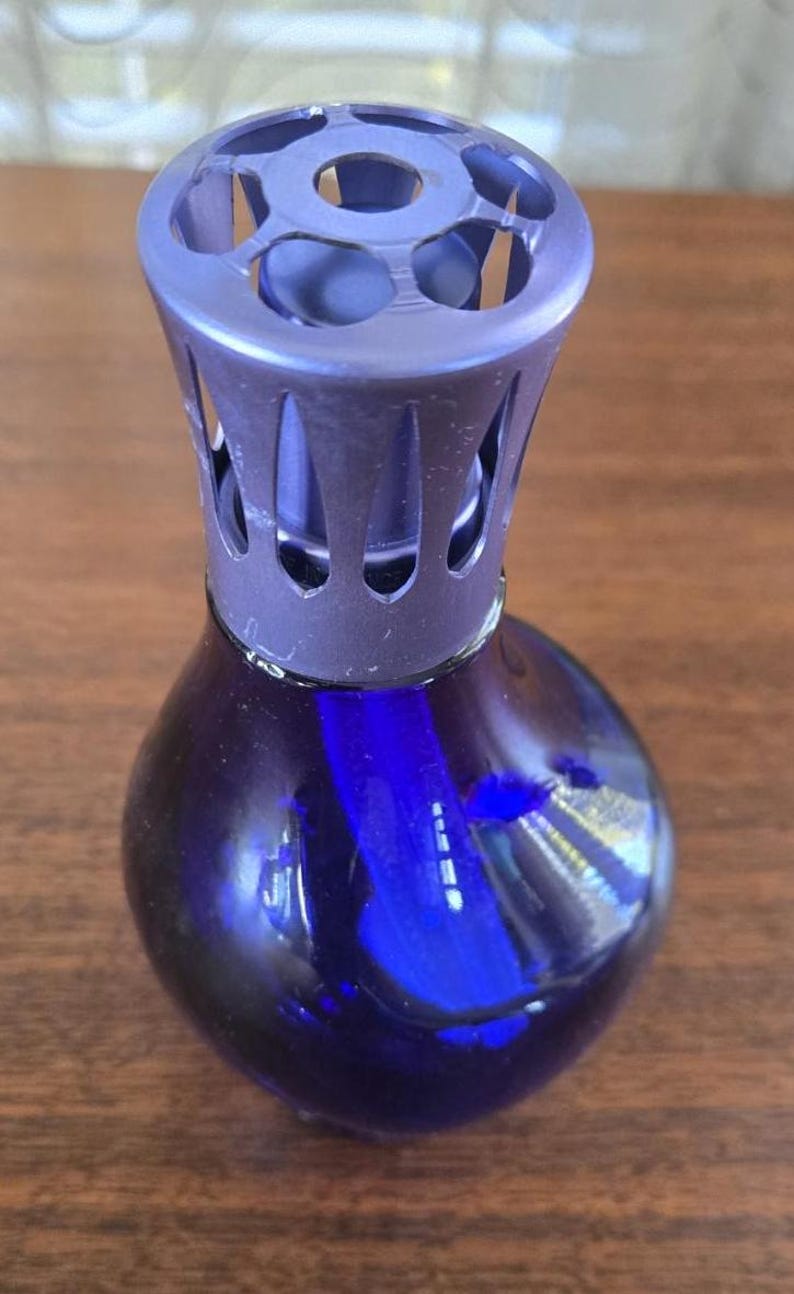 Pu&ograve; includere: Una bottiglia diffusore di fragranze in vetro blu scuro con un tappo decorativo, traforato, blu-viola. La bottiglia ha una forma arrotondata e una superficie liscia e riflettente. Il tappo ha un design geometrico.