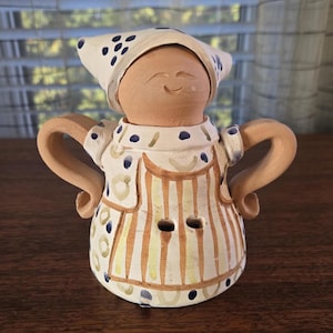 May include: A ceramic figurine of a woman wearing a blue and white patterned dress and a blue and white patterned hat. The figurine has a smiling face and is holding her arms out.