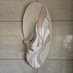 Peut inclure: Sculpture murale ovale texturée représentant un visage stylisé aux yeux fermés et aux lèvres entrouvertes. Le design intègre des accents roses, blancs et gris, avec des lignes fluides et une esthétique délicate. De fins brins s'étendent du visage.