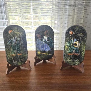 May include: Three oval-shaped decorative plaques featuring dark, speckled backgrounds with colorful, stylized figures. Each plaque is supported by a small, brown wooden stand. The figures are depicted in shades of blue, green, and orange.