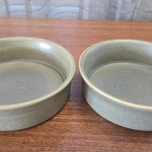 Puede incluir: Dos cuencos de cerámica verde oliva con forma ligeramente ovalada. Los cuencos tienen un acabado liso y brillante y un anillo azul oscuro alrededor de la base interior. Se colocan sobre una superficie de madera marrón.
