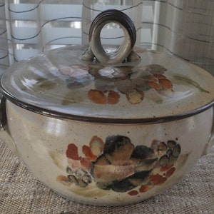 May include: A ceramic casserole dish with a lid, featuring a hand-painted floral design in shades of brown, orange, and green. The dish has two handles on the sides and a knob on the lid.