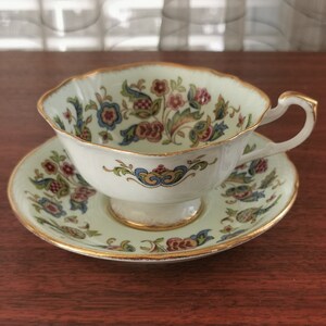 Può includere: Una tazza da tè e un piattino bianchi con sfondo verde e un motivo floreale in tonalità blu, rosse e dorate. La tazza ha un bordo ondulato e un bordo dorato.