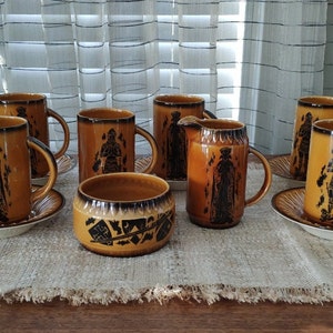 Puede incluir: Un juego de tazas, platillos y una jarra de cerámica vintage. Las tazas y la jarra son de color marrón cálido con ilustraciones negras de figuras. Los platillos son blancos con un borde marrón. El juego está dispuesto sobre una superficie texturizada de color neutro.