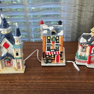 Op de afbeelding: Drie keramische kerstdorpgebouwen met witte stroomkabels. Het linkergebouw is een kerk met een blauw dak en een rood kruis. Het middelste gebouw is een winkel met meerdere verdiepingen en een rode deur. Rechts staat een school met een klokkentoren.