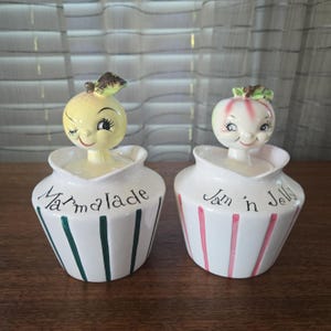 Puede incluir: Un par de tarros de cerámica vintage con tapas en forma de fruta. Uno está etiquetado como "Marmalade" y tiene rayas verdes, mientras que el otro dice "Jam n Jelly" con rayas rosas. Las tapas son personajes de frutas sonrientes.