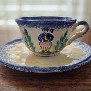 May include: A vintage teacup and saucer set with a hand-painted design of a person in traditional clothing. The cup and saucer are white with blue, green, yellow, and pink accents.