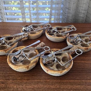 May include: Six ceramic escargot dishes with stainless steel tongs and forks. Each dish has six indentations for the snails. The dishes are brown with a speckled glaze and sit on a wooden surface.