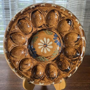 Puede incluir: Una fuente de cerámica marrón para servir huevos con 12 hendiduras en forma de huevo alrededor del perímetro y un pozo central. La fuente presenta un diseño floral en el centro y un borde decorativo.