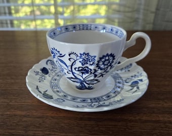 May include: White teacup and saucer set with blue floral patterns. The teacup has a unique, faceted shape and a curved handle. The saucer has a scalloped edge. The design features detailed blue floral motifs on a white background. A classic tea set.