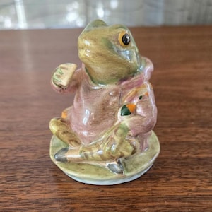 Op de afbeelding: Een keramieken beeldje van een kikker met een roze jas en een klein voorwerp. De kikker is groen en bruin met gele ogen. Hij zit op een groen en bruin voetstuk. Het beeldje is waarschijnlijk een decoratief item.