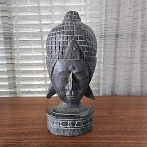 Puede incluir: Una escultura de madera gris oscuro de una cabeza de Buda. La cabeza tiene un patrón detallado, similar a una cuadrícula, en la parte superior y una expresión facial serena. La escultura se asienta sobre una base decorativa.