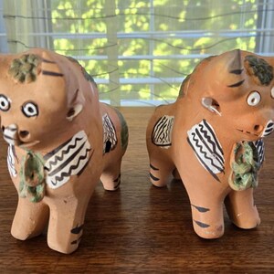 Puede incluir: Dos figuras de toro de cerámica marrón con detalles pintados en verde, blanco y negro. Los toros tienen ojos grandes y están de pie sobre una superficie de madera.