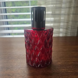 Puede incluir: Una lámpara de fragancia cilíndrica de vidrio rojo con un patrón de textura en forma de diamante en toda su superficie. La lámpara cuenta con una tapa de metal negro ventilada en la parte superior, ajustada al cuello del recipiente de vidrio.