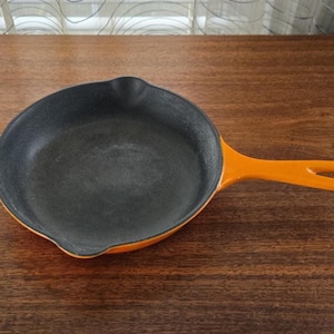 Pode incluir: Uma frigideira de ferro fundido laranja com interior preto. A frigideira tem uma alça longa com um furo para pendurar. A panela está sobre uma superfície de madeira marrom escura.