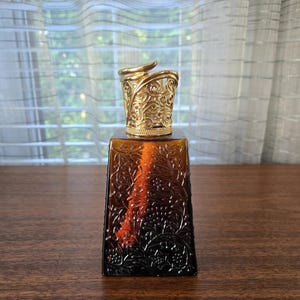 May include: A square-shaped amber glass fragrance lamp with a dark brown gradient and an intricate embossed floral pattern. The top features a decorative gold-toned metal crown with a swirling filigree design, sitting atop the glass vessel.